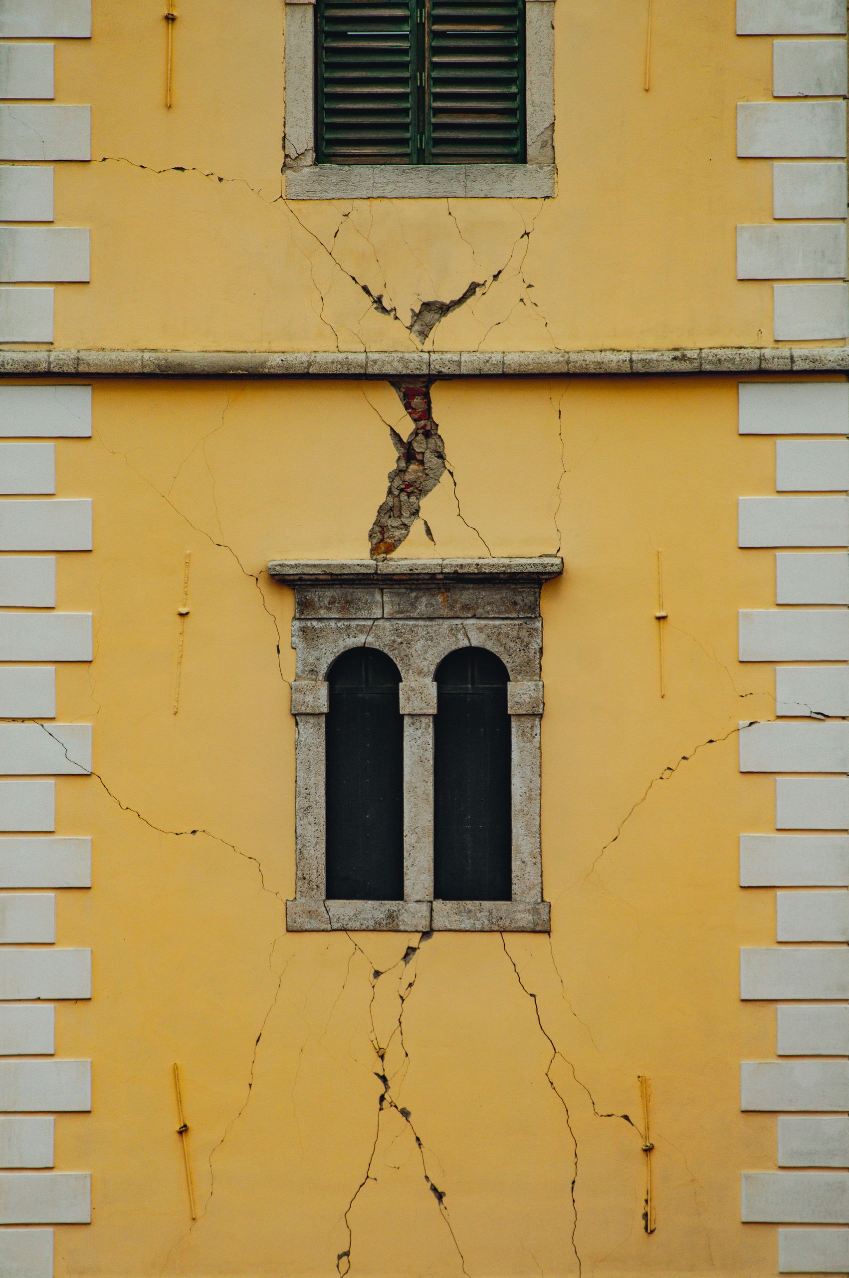 Close-up of a weathered yellow building façade with cracks, featuring arched windows and shutters.